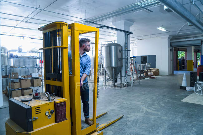 Молодой человек на пивоваренном заводе водит вилочный погрузчик, смотрит в сторону — стоковое фото