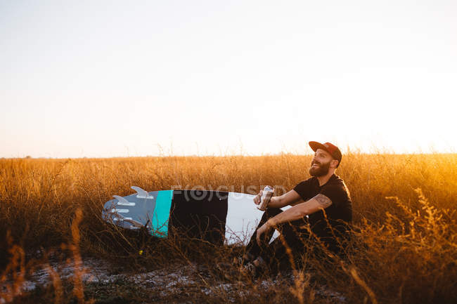 Männlicher Surfer trinkt Bier im Feld aus langem Gras bei Sonnenuntergang, San Luis Obispo, Kalifornien, Vereinigte Staaten — Stockfoto