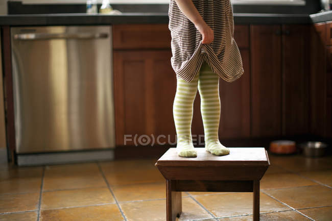 Recorte de piernas de niño de pie en el taburete en la cocina - foto de stock