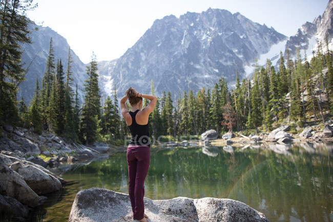 Young woman standing on rock beside lake, looking at view, The Enchantments, Alpine Lakes Wilderness, Washington, USA — Stock Photo