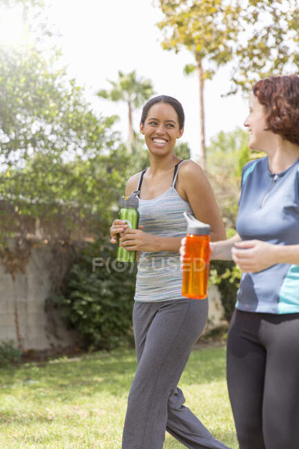Mujeres jóvenes caminando usando ropa deportiva llevando botellas de agua riendo - foto de stock