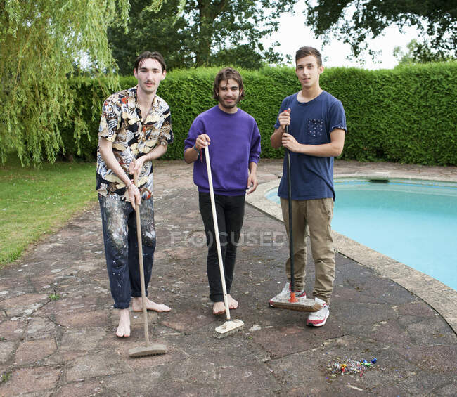 Portrait of three young men on poolside with brooms — Stock Photo