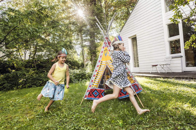 Two girls in native American headdress running around teepee in garden — Stock Photo
