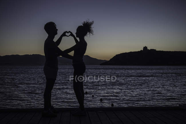 Couple faisant forme de coeur avec les mains par la mer, silhouette — Photo de stock