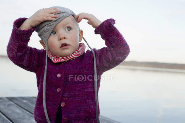 Baby girl on lake pier trying to put on hat — Stock Photo