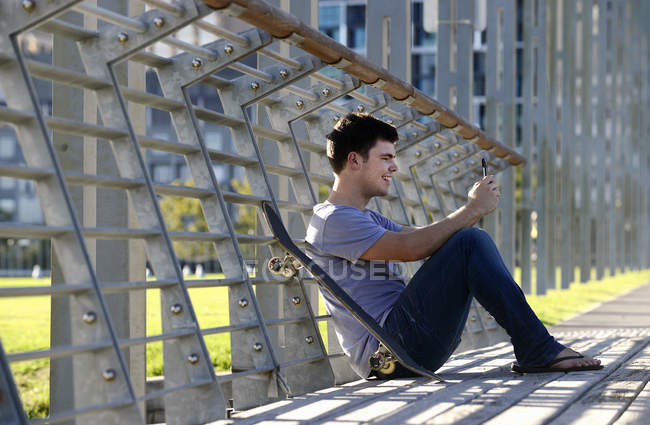 Joven sentado, usando teléfono móvil, monopatín al lado - foto de stock