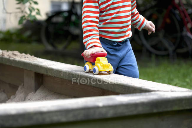 Tiro cortado de criança masculina empurrando carro de brinquedo em torno do poço de areia no jardim — Fotografia de Stock