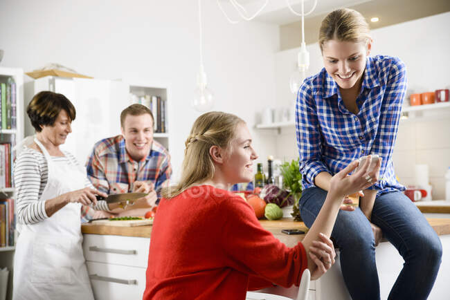 Due donne con cellulare in cucina, madre e fratello in background — Foto stock