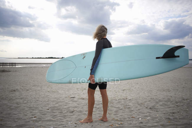 Seniorin steht am Strand, hält Surfbrett, Rückansicht — Stockfoto