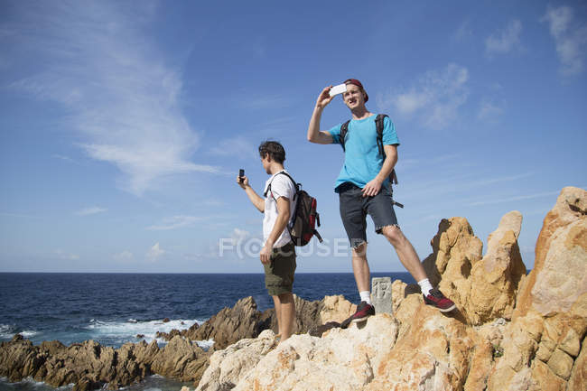 Jóvenes parados sobre rocas usando smartphone para fotografiar, Costa Paradiso, Cerdeña, Italia - foto de stock