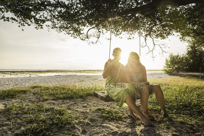 Dos mujeres sentadas en el columpio del árbol de playa al atardecer, Gili Trawangan, Lombok, Indonesia - foto de stock