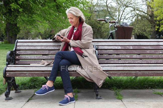Donna anziana seduta sulla panchina del parco e guardando il telefono cellulare — Foto stock