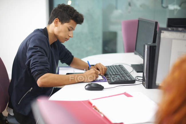 Молодий студент коледжу за комп'ютерним столом робить нотатки — стокове фото