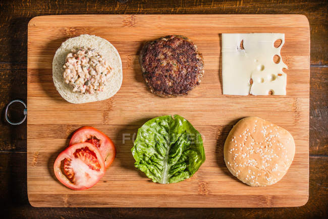 Chopping board with hamburger and condiments — Stock Photo