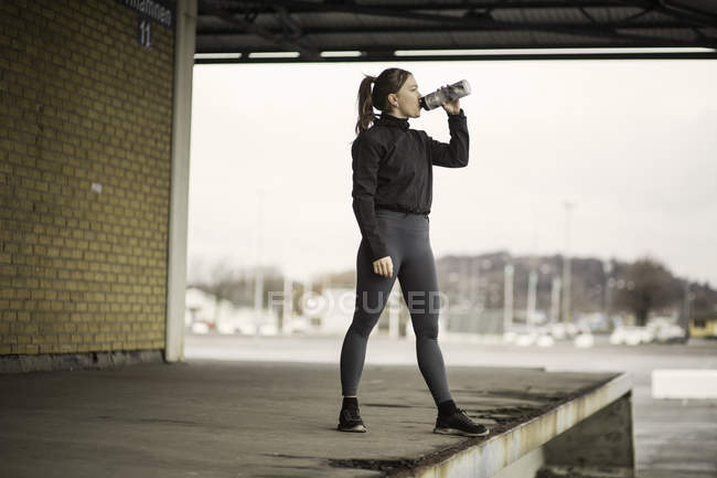 Female runner drinking bottled water on warehouse platform — Stock Photo