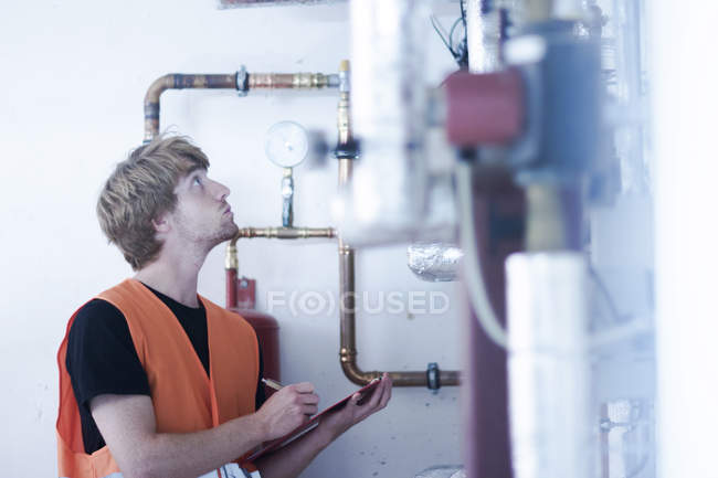 Jeune homme dans la chaufferie tenant le presse-papiers inspectant le système de chauffage, regardant vers le haut — Photo de stock