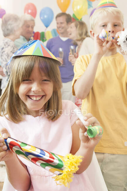 Niño y niña usando sombreros de fiesta mirando a la cámara soplando cuernos de fiesta sonriendo - foto de stock