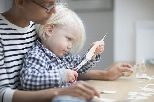 Niño usando tijeras para cortar papel con su madre - foto de stock