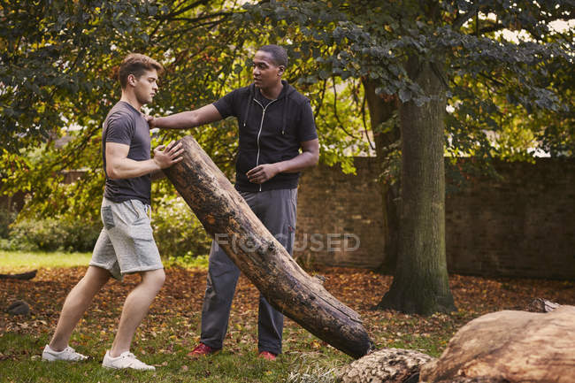 Jeune homme avec entraîneur personnel soulevant tronc d'arbre dans le parc — Photo de stock