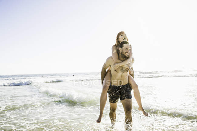 Man wearing swimming shorts giving girlfriend piggyback in sea, Cape Town, South Africa — Stock Photo