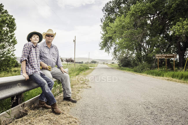Agricultor y nieto adolescente sentado en la barrera de la carretera - foto de stock
