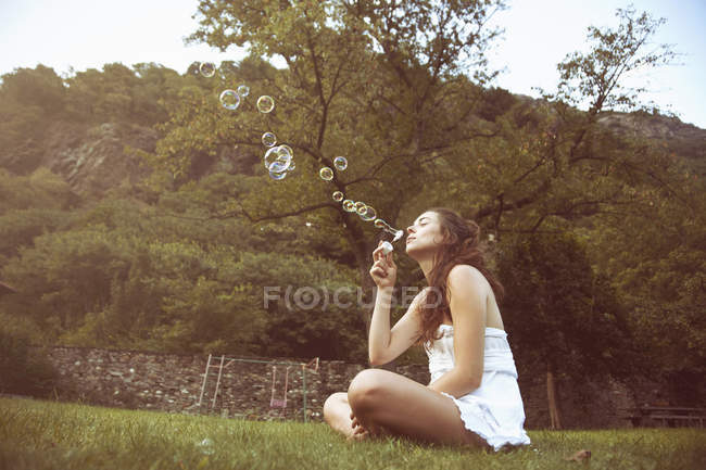Young woman sitting cross legged in field blowing bubbles — Stock Photo