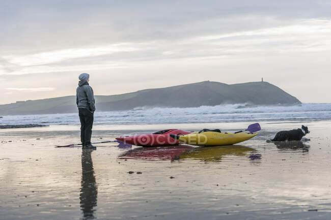 Mujer joven en la playa con kayaks de mar, Polzeath, Cornwall, Inglaterra - foto de stock