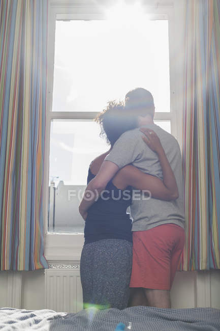 Mid adult couple, hugging, looking out of window, rear view — Stock Photo