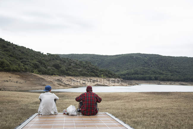 Вид сзади на молодых людей, сидящих на деревянном пирсе, смотрящих на горный хребет, Нуоро, Сардиния, Италия — стоковое фото