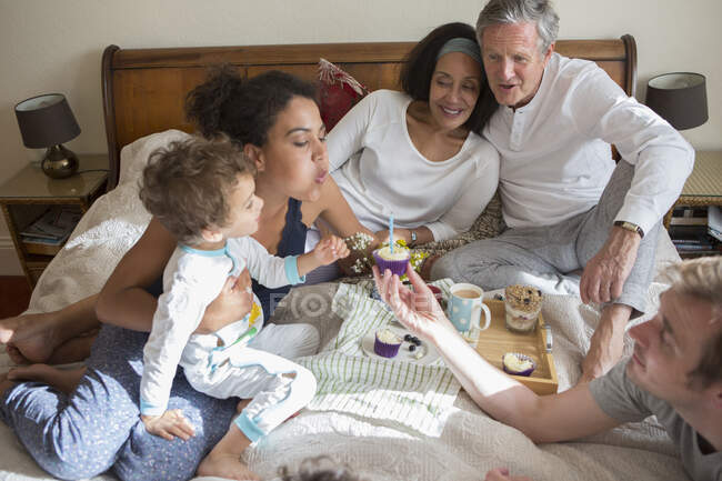 Família de três gerações sentada na cama, soprando vela no cupcake — Fotografia de Stock