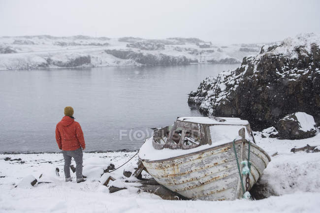 Hombre por lago con barco en paisaje nevado, Islandia - foto de stock