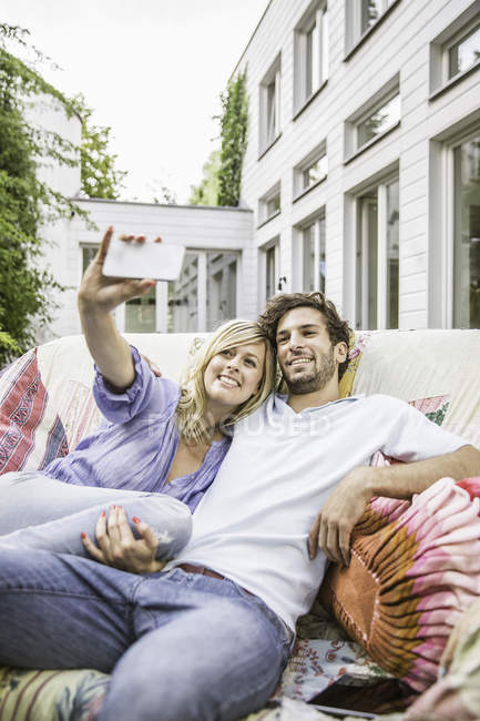 Пара сидящих на винтажном диване в саду и делающих селфи на смартфоне — стоковое фото