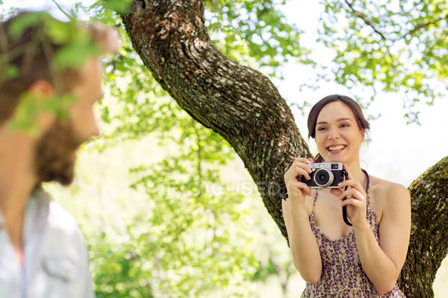 Молодая женщина под деревом фотографирует молодого человека — стоковое фото