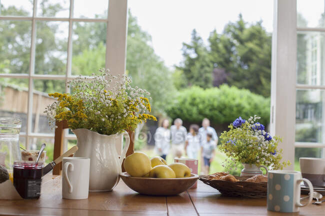 Mesa de cocina con alimentos para el desayuno, familia en el jardín detrás, se centran en la mesa - foto de stock