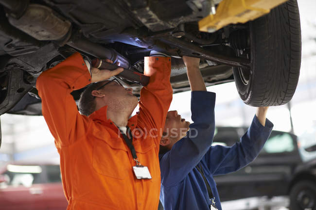 Estudiantes de mecánica universitaria inspeccionando debajo del coche en el garaje de reparación - foto de stock