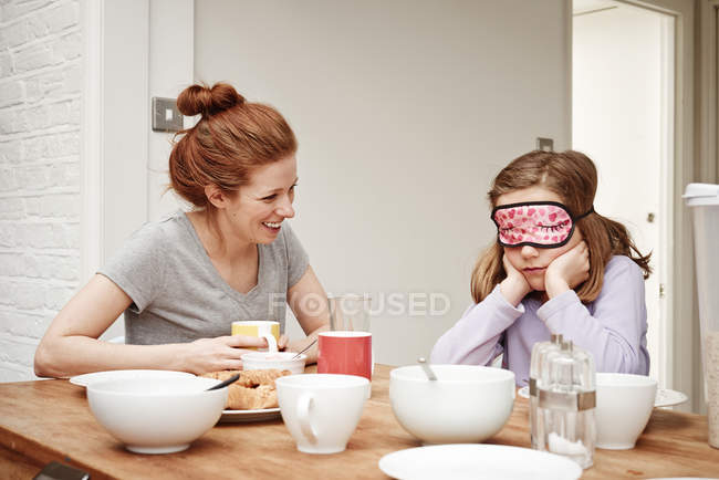 Mulher adulta média na mesa de café da manhã com a filha usando máscara de sono — Fotografia de Stock