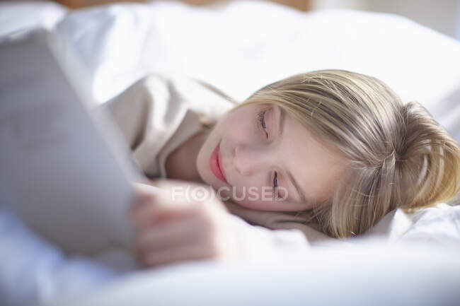 Blond haired girl lying in bed reading a book — Stock Photo