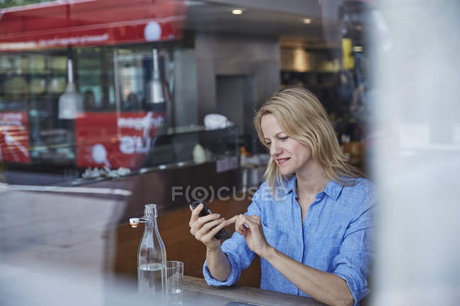 Mulher madura sentada no café, usando smartphone, ônibus refletido na janela — Fotografia de Stock