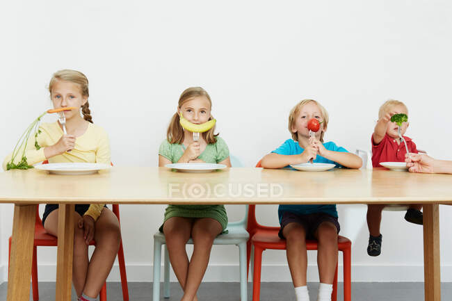 Cuatro niños sentados a la mesa con verduras en tenedores - foto de stock