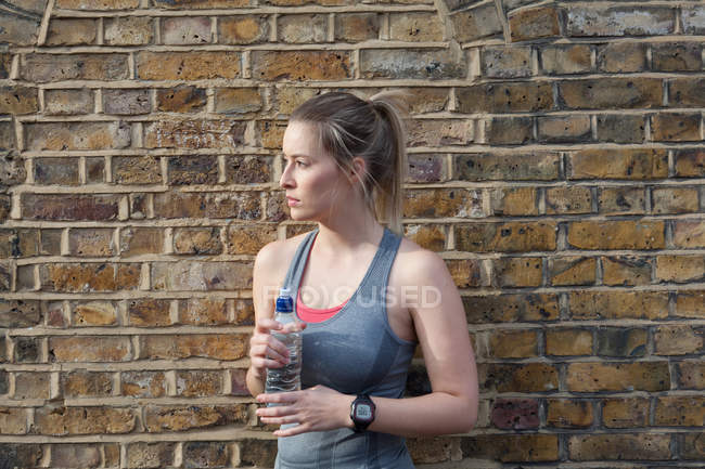 Молодая бегунья перед кирпичной стеной пьет воду — стоковое фото