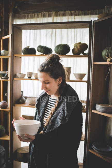 Молода жінка тримає керамічну тарілку перед полицями, показуючи глиняні горщики та гарбузи — стокове фото