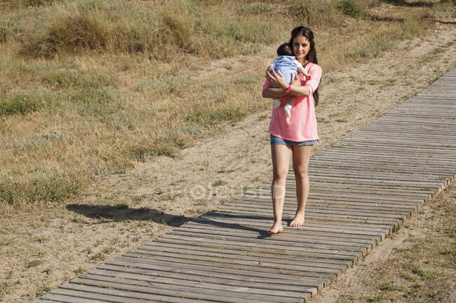 Madre caminando a lo largo del paseo marítimo llevando al bebé en brazos - foto de stock