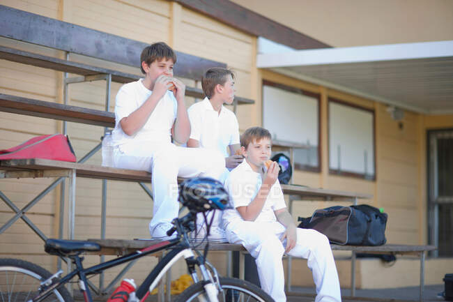 Tres chicos sentados en gradas con ropa de cricket - foto de stock