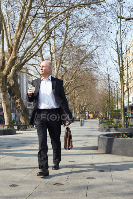 Зрелый бизнесмен ходит по тротуару с кофе — стоковое фото