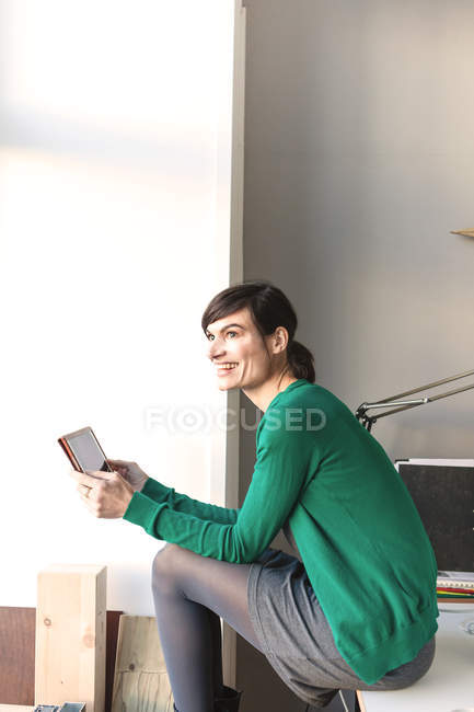 Vista lateral de la mujer madura sentada en el escritorio sosteniendo la tableta digital mirando hacia otro lado sonriendo - foto de stock
