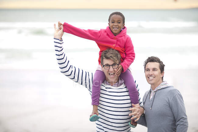 Гей-пара, гуляющая по пляжу с ребенком на плечах — стоковое фото