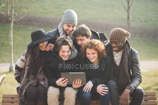Seis jovens amigos adultos usando tablet digital no banco do parque — Fotografia de Stock