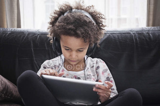 Menina idade elementar sentado no sofá usando tablet digital e fones de ouvido — Fotografia de Stock
