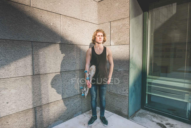 Retrato de un joven skateboarder urbano de pie en la esquina sosteniendo el monopatín - foto de stock