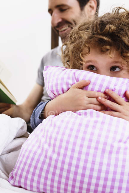 Chica abrazando almohada mientras padre leyendo libro de cuentos en la cama - foto de stock